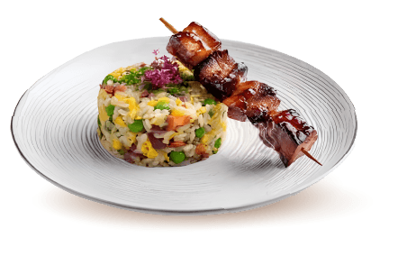 A gourmet entrée featuring a beautifully molded round stack of seasoned fried rice mixed with egg and green peas, served alongside a glistening, teriyaki-glazed grilled meat skewer on a textured white ceramic plate.